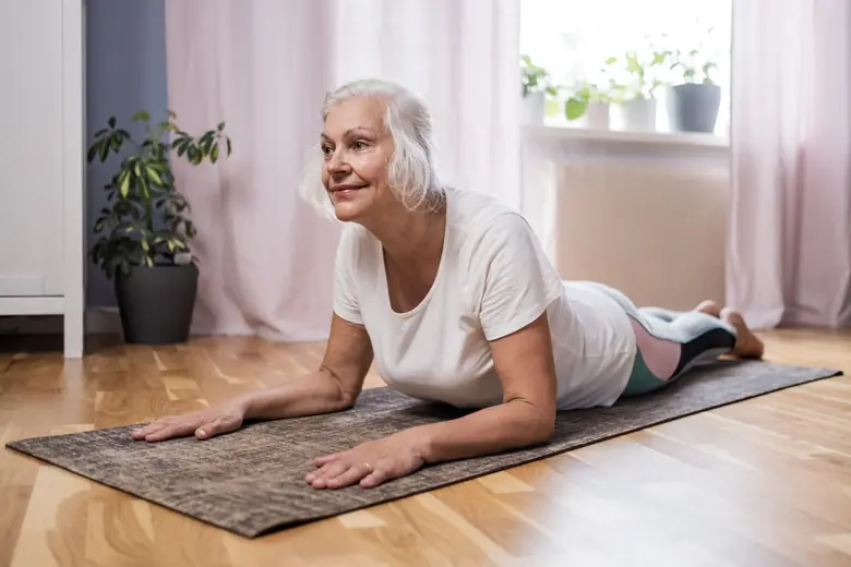 Mulher de aproximadamente 50 anos fazendo exercício de alongamento no chão da sala de casa, com luz natural e expressão concentrada