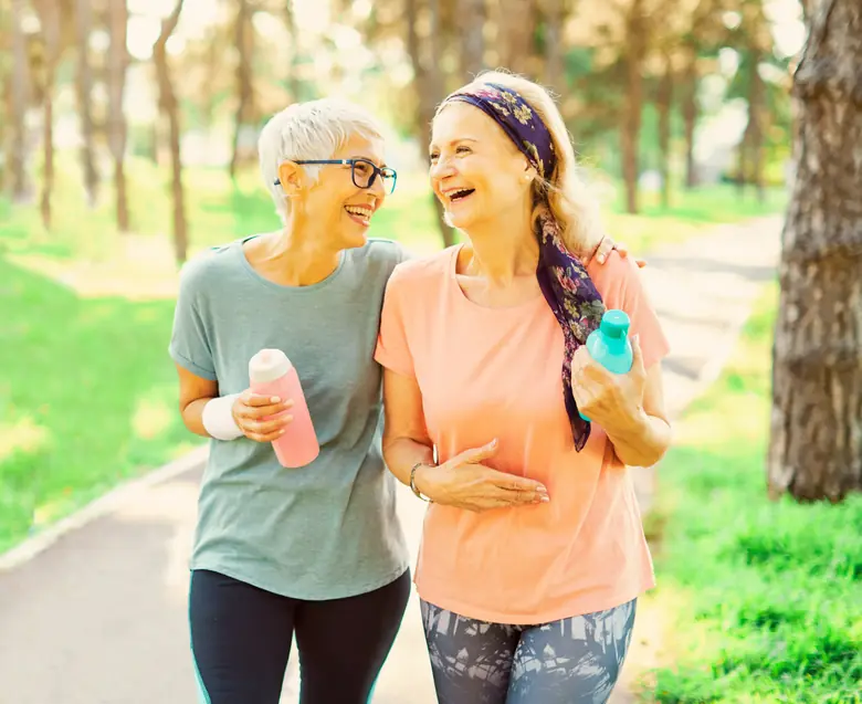 Duas mulheres por volta dos 50 anos caminhando e rindo em um parque ao ar livre, com aparência saudável e ativa
